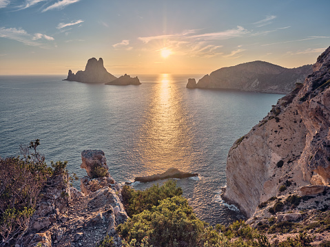 Wide-angle view of the sun setting over the strait between the iconic isles of Es Vedrà and Es Vedranell (left), and the rough strip of land of Puig des Savinar (right). Blue sky, picturesque clouds, calm waters reflecting the oblique, golden light of a Mediterranean summer sunset, steep cliffs dotted with Mediterranean scrub and trees. Photo taken from the southernmost point of Cap Llentrisca. The small rock known as Galereta de Cala Llentrisca can be seen at the bottom. High level of detail, natural rendition, realistic feel. Developed from RAW.