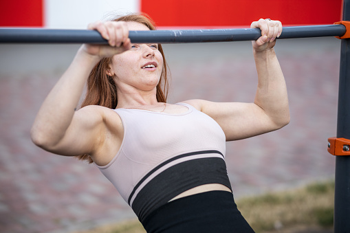 red-haired girl pulls up on the horizontal bar outdoors, doing sports outdoors on the sports ground. strong girl