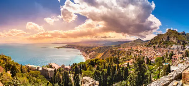 Photo of Taormina, Sicily, Italy. Panoramic view over Taormina town on hilltop, coastline of scenic bay in Ionian sea, Naxos town and mountains unter sunset sky. Popular tourist destination