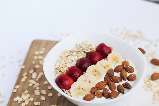Healthy fruit salad with almonds, bananas, cherries, cornflakes and yogurt. Served in a white bowl on a wooden board. Kitchen table with white sofa.