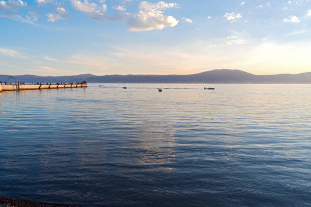 Chapala, JALISCO/MEXICO - March 4, 2023: Walkers along pier viewing lake at sundown and boats on water with mountains behind them.