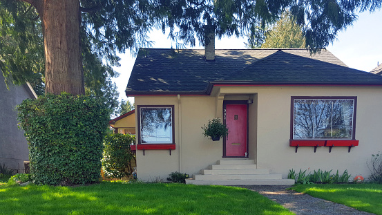 New Westminster, British Columbia, Canada- April 29,2023: Exterior front view of bungalow built in 1944 just before the end of World War 2. 
Beautiful condition. Giant cedar tree in front yard. Lawn,sidewalk up to front stairs.  Red door and trim.