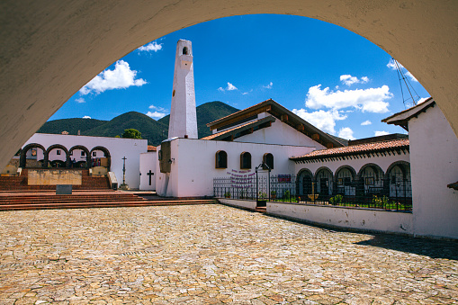 Guatavita, Colombia - The view from under an arched entrance to the main town square in Guatavita, in the Cundinamarca department of the South American country of Colombia. The church on the square with its bell tower can be seen; and to the left, the principal entrance to the square. The altitude is about 8,660 feet above mean sea level. In the background are the Andes mountains. Photo shot in the afternoon sunlight;  horizontal format, no people. Copy space. Note to Inspector: on the wall of the church is a prayer. It is not an advertisement.