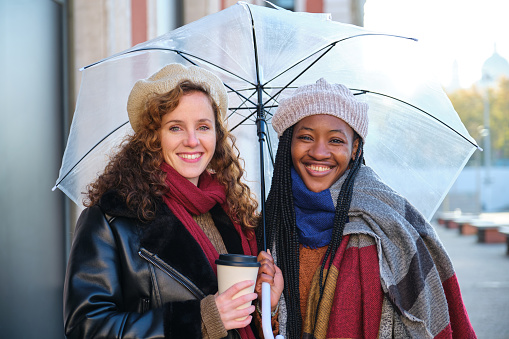 Two multiracial female friends looking at camera and smiling in winter, holding an umbrella and a coffee cup.