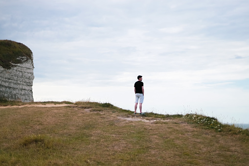 One young caucasian brunette guy in a black T-shirt and light shorts stands from the back on a mountain top on a cloudy day admiring the view of the northern sea, close-up side view.