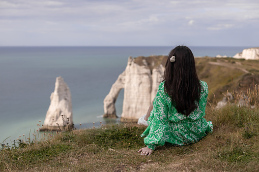 One young caucasian brunette girl in a green dress with flowing hair sits from the back on a mountain top enjoying the view of the northern sea and a natural arch on a gloomy spring day in Etretat, close-up side view.