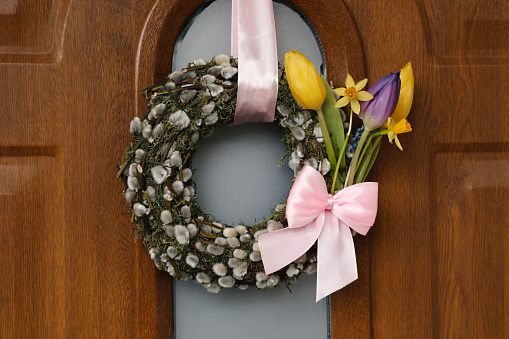 Wreath made of beautiful willow branches, pink bow and colorful tulip flowers on wooden door
