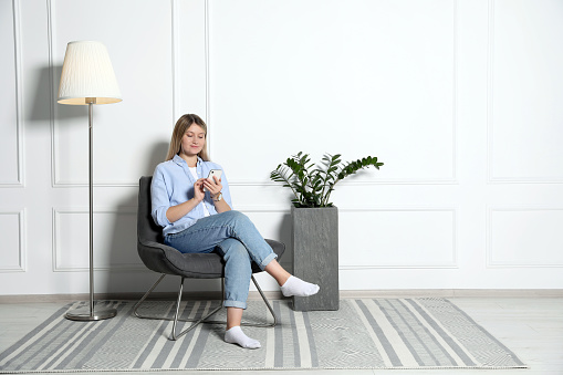 Young woman with phone sitting in armchair at home, space for text. Interior design