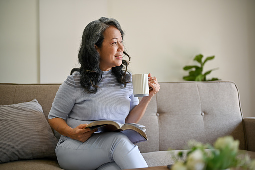 A happy and relaxed retired Asian woman is looking out the window and daydreaming about her happy life while relaxing on a sofa in her living room.