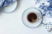 White cup of coffee with saucer, blue and white napkins  on a white table. View from above.