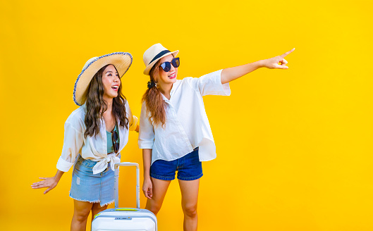 Two Pretty asian women passengers in trendy fashion is exciting to board while carrying her luggage bag in happiness for friendly trip and summer travel vacation isolated on yellow background