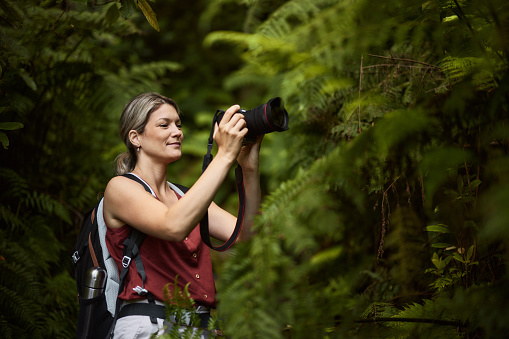 Smiling female backpacker using DSLR camera while taking a photo among greenery in nature.