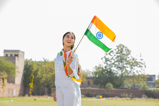 Young beautiful woman wearing traditional white dress holding indian and weaving flag while standing at park celebrating Independence day or Republic day.