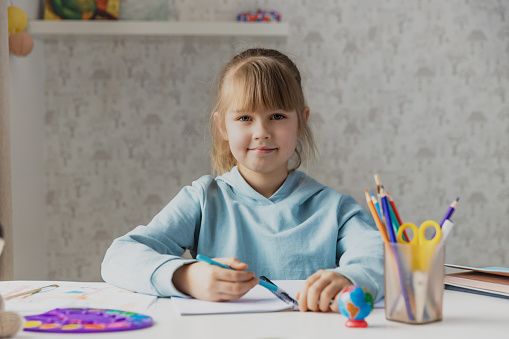 Schooler cute little girl sitting at desk in bedroom, holding a pen, doing homework, kid studying at home, getting ready before exams, writing notes. Home education, homeschooling. Child is happy