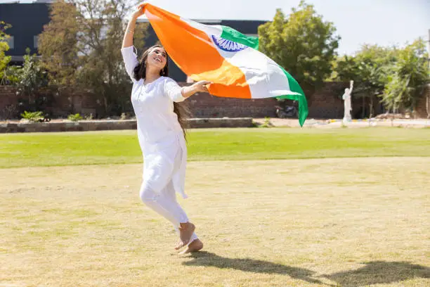 Happy young woman wearing traditional white dress running with big indian flag outdoor at park, celebrating Independence day or Republic day.