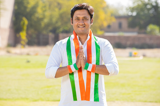 Portrait of young indian man wearing traditional white kurta and tricolor duppata do namaste while standing at park. Election and politics, celebrating Independence day or Republic day.