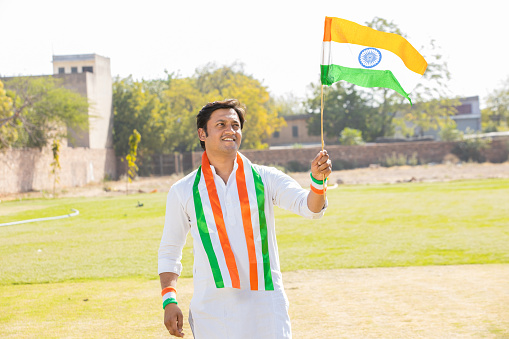 happy young man wearing traditional white dress holding indian and weaving flag while standing at park celebrating Independence day or Republic day.