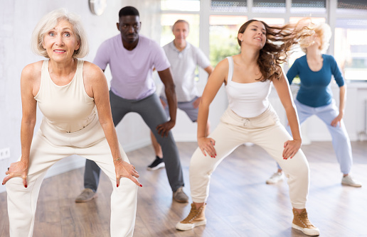 Cheerful active senior woman having fun and dancing hip hop during workout in dance class with mixed age multiracial group of adults