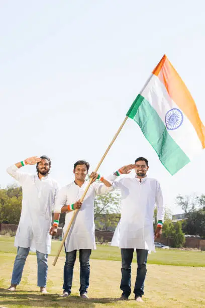 Group of happy young men wearing traditional white dress holding indian flag and saluting, celebrating Independence day or Republic day.