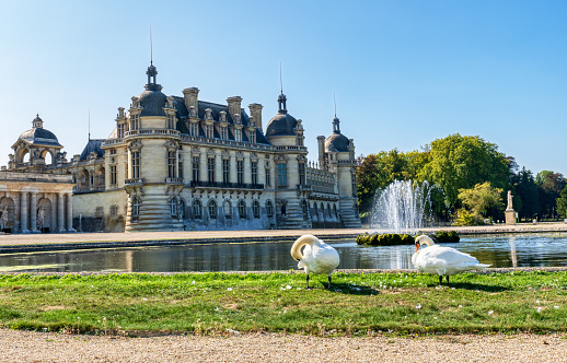 Chantilly, France - September 21 2020: View of the Chateau de Chantilly from Le Notre Garden with a fountain and two swans in the foreground - France