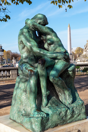 Paris, France - November 22 2022: Bronze statue The Kiss (1882) by Auguste Rodin (1840-1917) in the Tuileries Garden, in front of the Orangerie museum, with Egyptian obelisk in background - Paris, France