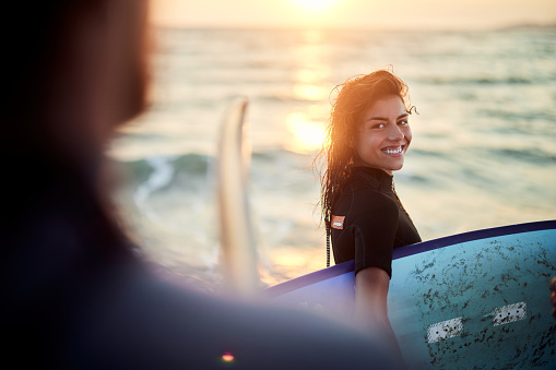 Happy female surfer carrying surfboard and communicating with her friend on the beach at sunset. Copy space.