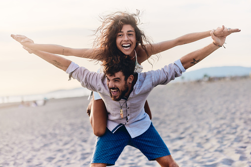Young playful couple having fun while piggybacking with their arms outstretched on the beach. Copy space.