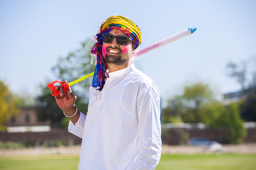 Young indian man with sunglasses holding pichkari in hand play holi festival with his face painted with powder color or gulal.