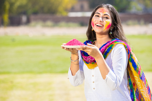 Portrait of smiling beautiful young Indian woman wearing white kurta dress holding pink powder color plate celebrating holi festival at park outdoor, Face painted with colorful gulal Copy space.