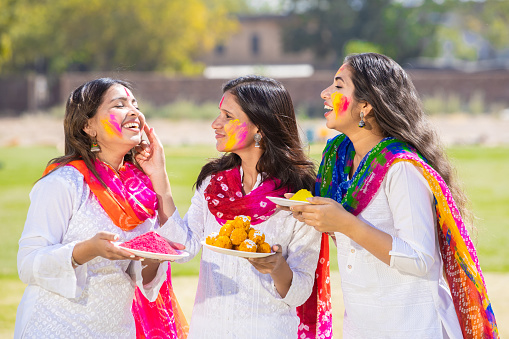 Group of happy young indian woman wearing white kurta dupatta outfit celebrating holi festival at park while holding plates full of gulal and laddoo sweet , woman having fun with colorful powder.