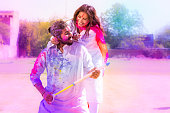 Cheerful young Indian couple playing with colorful powder color or gulal celebrating holi festival at park outdoor.