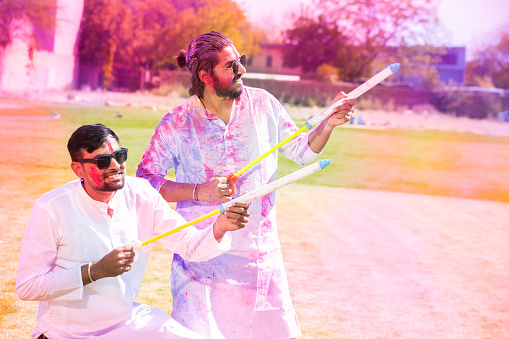 Young Indian people wearing white kurta holding pichkari celebrating holi festival at park outdoor, Two Adult male friends Face painted with colorful powder color or gulal