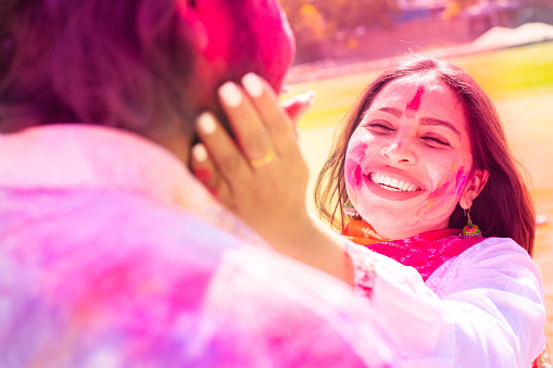 Happy young Indian couple having fun playing with colorful powder color or gulal celebrating holi festival at park outdoor. Closeup.