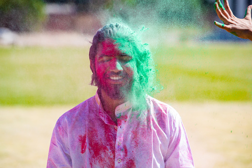 Portrait of happy Indian man wearing sunglasses and painted face celebrating Holi festival with colorful powder or gulal. Closeup.