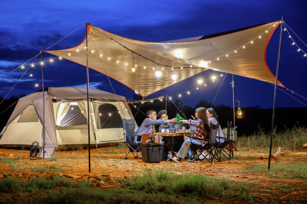 Outdoor camping tent in the forest park, party dinner with friends under tarp or flysheet and warm lighting at night near natural under a blue twilight sky. Overview of travel camping with friends. tienda de campaña al aire libre en el parque forestal, cena de fiesta con amigos bajo lona o flysheet y una iluminación cálida por la noche cerca de natural bajo un cielo azul crepuscular. descripción general de los viajes de camping con amigos.
