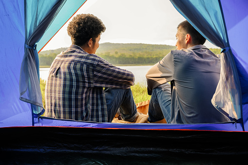 Young asian man sits and talk in front of the camping tent with natural mountain view and lake in background.