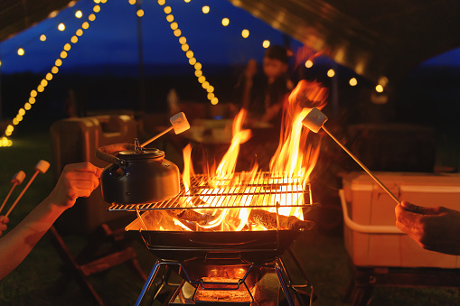 Roasting marshmallows on campfire grill in romantic night camping yard and night light with twilight sky blur background. Dessert food camping for child with nature outdoor, Focusing on marshmallows  **Noise**