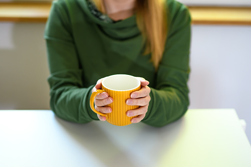A woman sitting at a table holding a yellow mug with a warm drink in both hands closeup