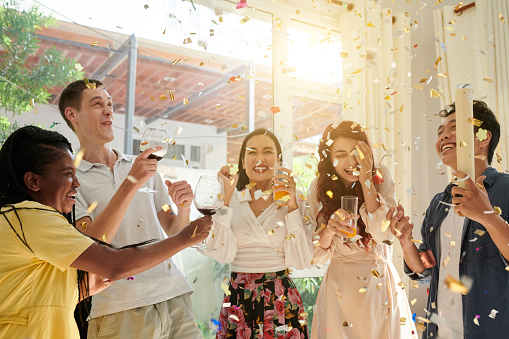 Group of excited friends dancing under falling confetti at home birthday party