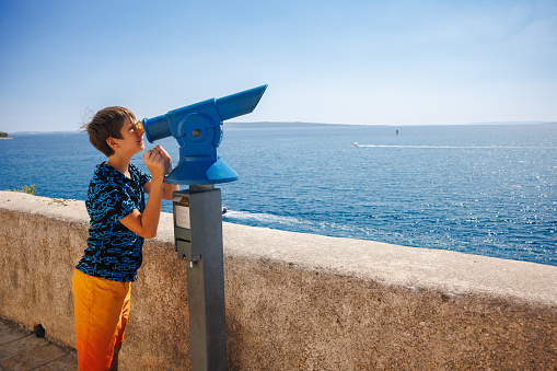 Preadolescent boy looking through binoculars from the town panoramic view platform in the town of Rab on Rab Island, sunny day in summer