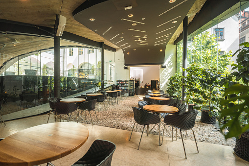 Wide angle shot of a fashionable brunch restaurant with  glass wall and  cool furniture. Modern design of cobblestone and wooden flooring. Daylight coming in. Coffee plant in the back of a coffee shop.