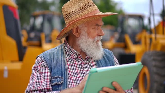Bearded confident senior man with digital tablet checking online database looking around at yellow tractors. Front view portrait of male Caucasian seller on market outdoors.
