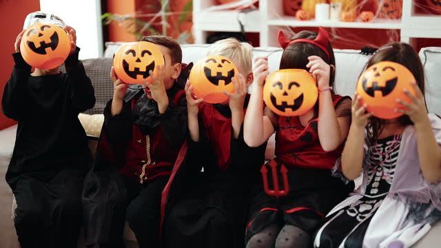 Group of kids wearing halloween costume covering face with pumpkin basket at home