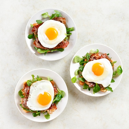 Open sandwiches with fried eggs, bacon and vegetable leaves on plate over light stone background. Top view, flat lay
