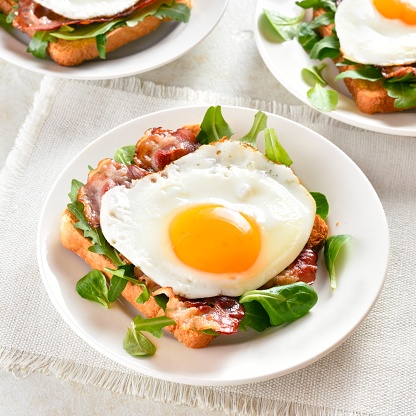 Open sandwiches with fried eggs, bacon and vegetable leaves on plate over light stone background. Close up view