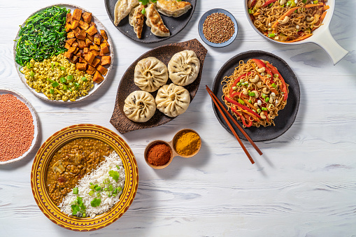 Vegan asian food assortment all plant based recipes as red lentils curry, gyozas, Stir fry Soba noodles, Steamed Panpao and Tofu salad scramble arrangement on white wood background