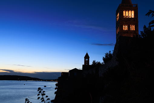 Beautiful view on illuminated church tower against dark blue sky at night, surrounding wall on Rab town waterfront, Rab island in Croatia