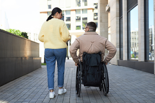 Rear view of young woman talking to her man with disability while they walking along the street in the city