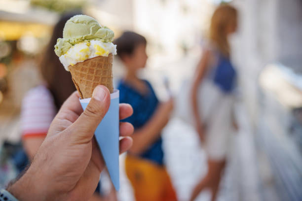 Holding a biscuit cone with pistachio and vanilla ice cream scoops, confectioner's display fridge for orders in the street