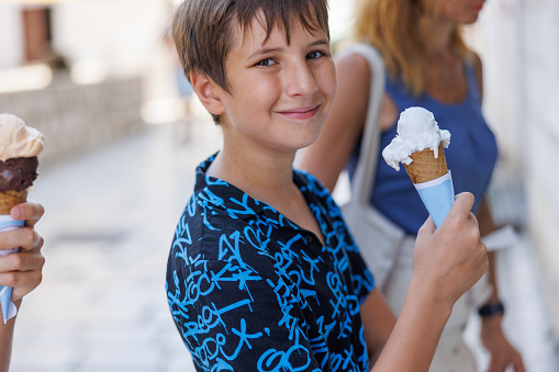 Preadolescent boy eating ice cream served on scoops in biscuit cone with his family in the street, smiling at camera